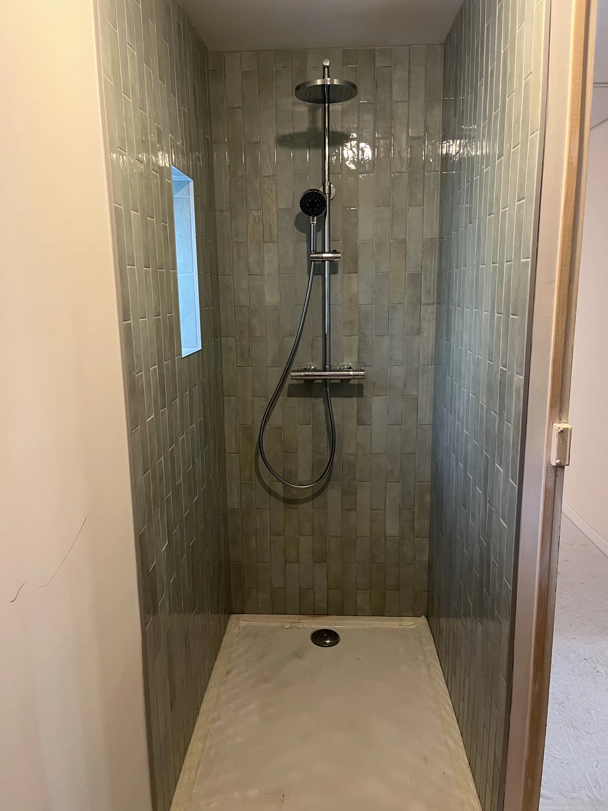 Douche avec carrelage mural vert, colonne chromée, douchette et pomme de tête au-dessus du receveur blanc.